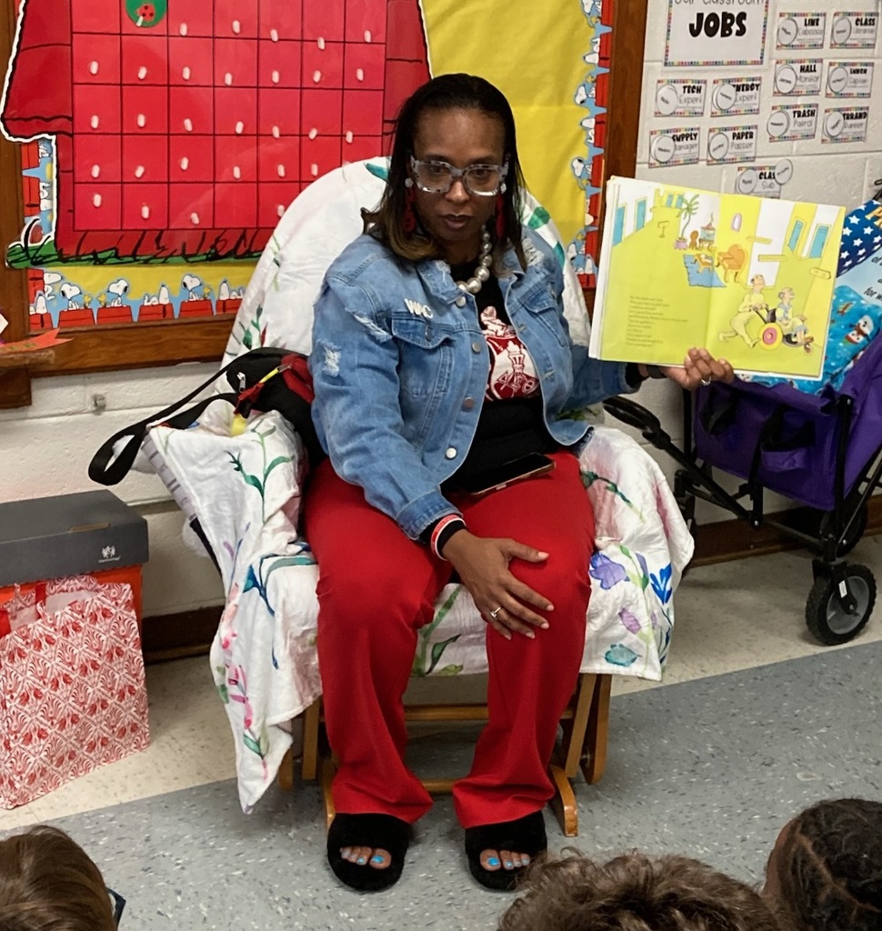 A teacher sits in a rocking chair in a colorful elementary classroom holding open a Dr. Seuss book while reading aloud. She is wearing red pants and a denim jacket. A bright bulletin board and classroom decorations are visible behind her.