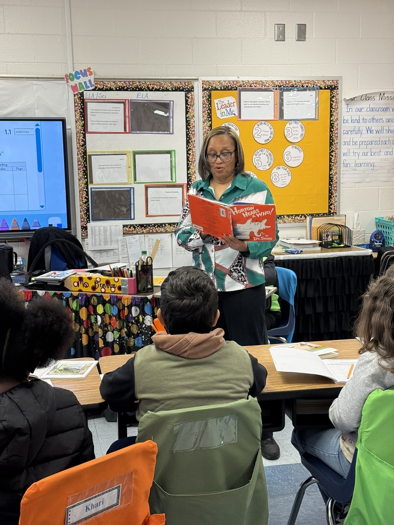 A teacher stands at the front of an elementary classroom reading Horton Hears a Who! by Dr. Seuss aloud to her students. Several students sit at their desks facing her and listening attentively. Behind the teacher are colorful bulletin boards, a “Leader in Me” display, and classroom learning targets posted on the wall.