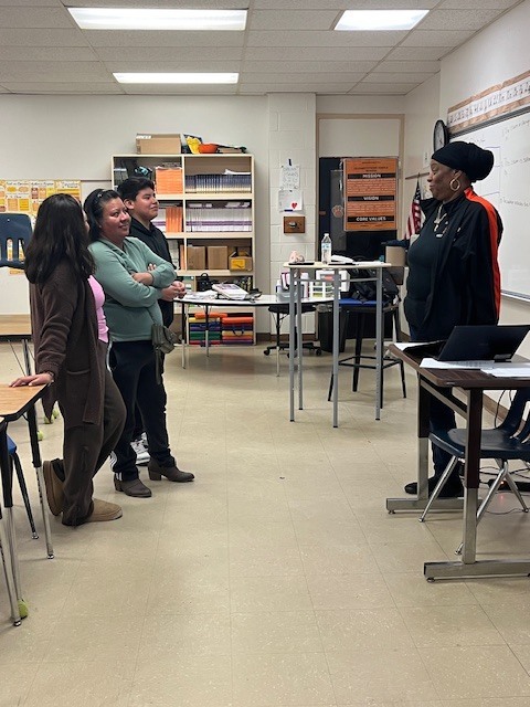Students stand at the front of a classroom speaking with a teacher while classmates watch, practicing communication and leadership skills.