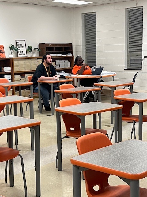 Two teachers sit at desks listening and preparing as students present during a classroom activity.