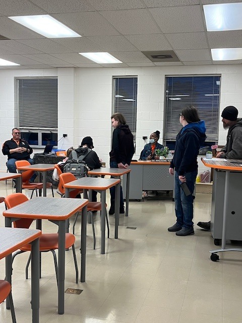 Students stand and present ideas to a teacher while classmates observe in a collaborative classroom discussion.