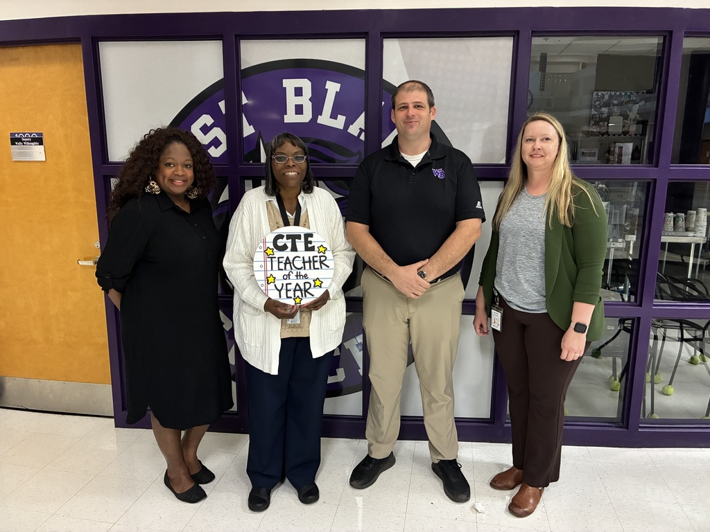 Four adults stand side by side in a school hallway in front of a purple-framed window featuring a large school logo. The person second from the left is holding a round sign that reads “CTE Teacher of the Year” with decorative stars. The group is dressed in business-casual attire and smiling at the camera. A classroom with tables and chairs is visible through the window behind them.