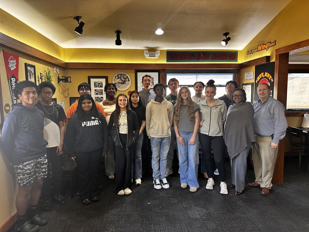 A group of high school students and two adults stand together inside a restaurant decorated with Pizza Hut memorabilia and signage reading “Elizabethtown’s Favorite Tailgating Zone.” The group faces the camera and poses for a photo.