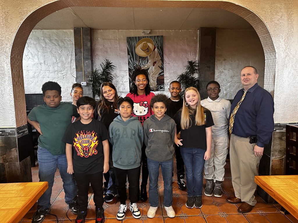 A group of elementary and middle school students stand with a school staff member inside a restaurant with tiled floors and an arched wall feature. The students smile and pose together for a group photo.