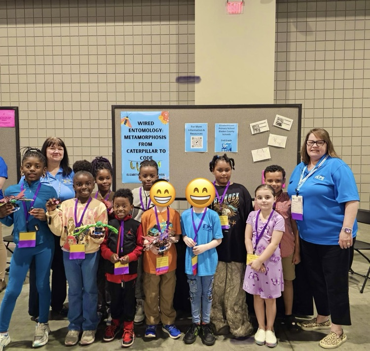 A group of Elizabethtown Primary School second grade students stand with their teachers at the NCTIES conference student showcase. The students wear conference badges and hold insect-inspired robotics projects they created.