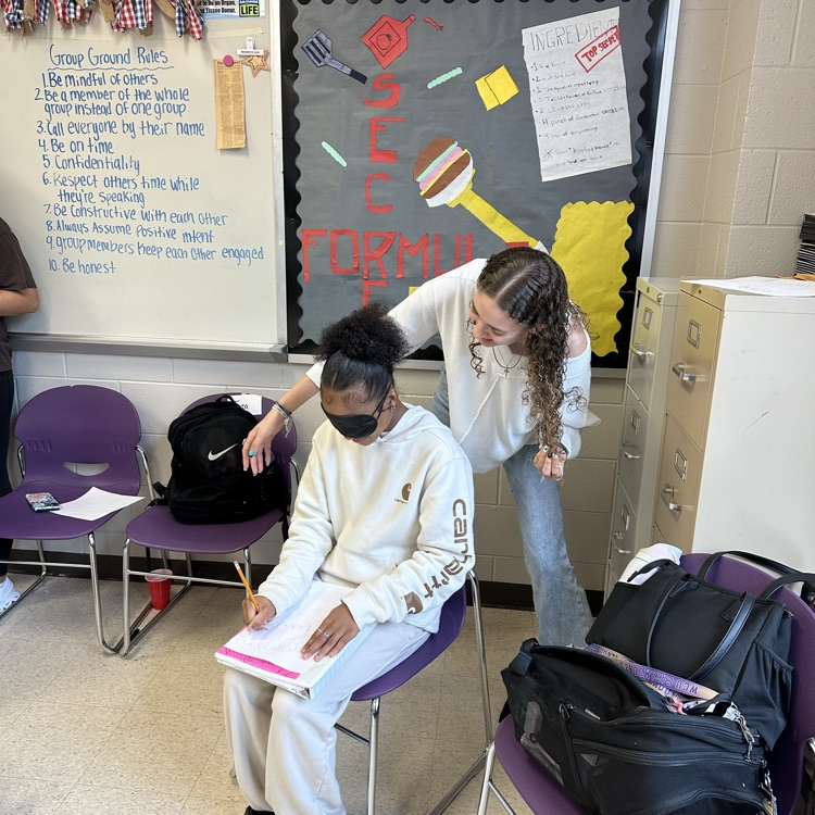 a blindfolded student sits in a chair while another student stands behind coaching her