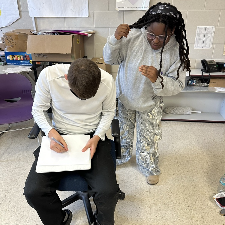 a blindfolded boy draws while a female student gives directions