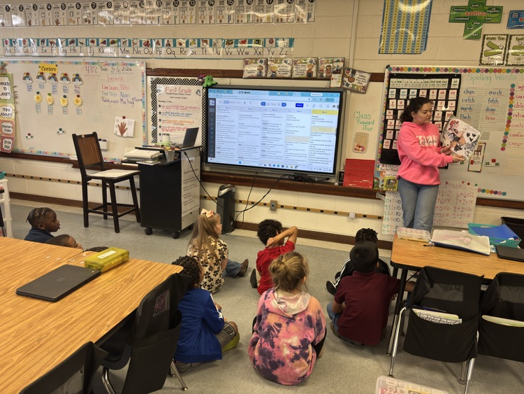 classroom full of small students sitting on the floor, listening to a story told by a East Bladen student in pink