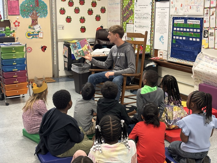 students crisscross on the floor, listening to a student in a gray shirt and a rocking chair story
