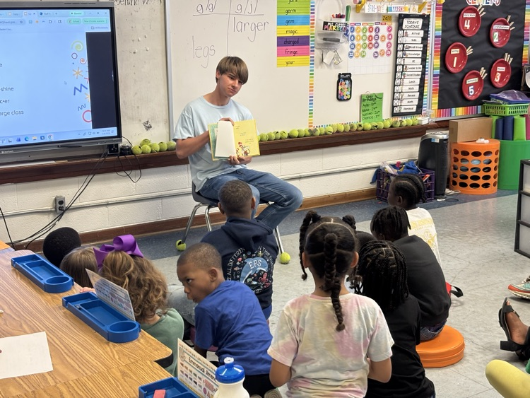 students crisscross on the floor, listening to a student in a light blue shirt and on achair story