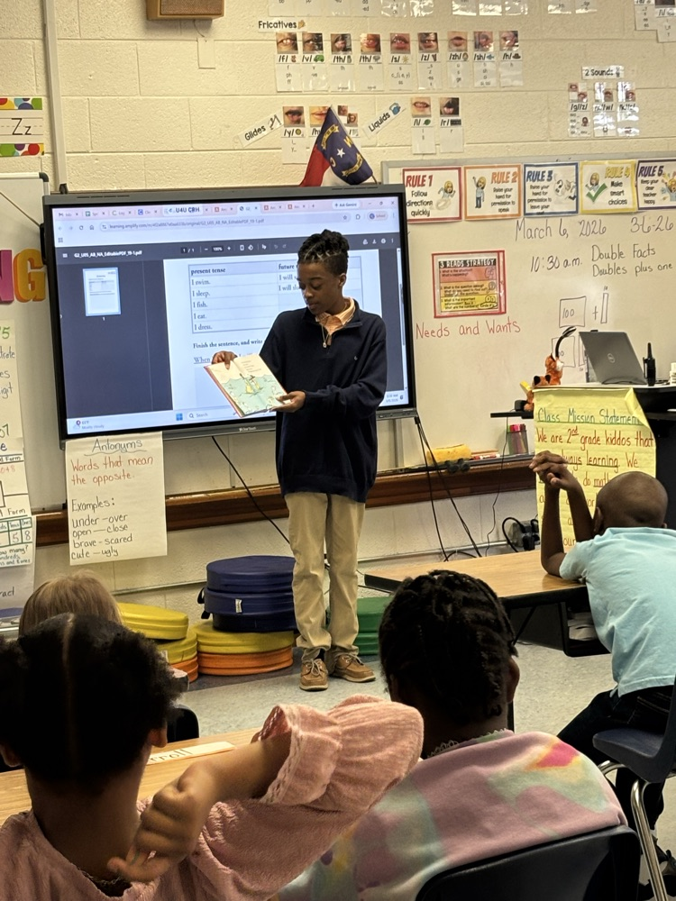 student sitting at their desk while a student in khaki pants reads a story to them