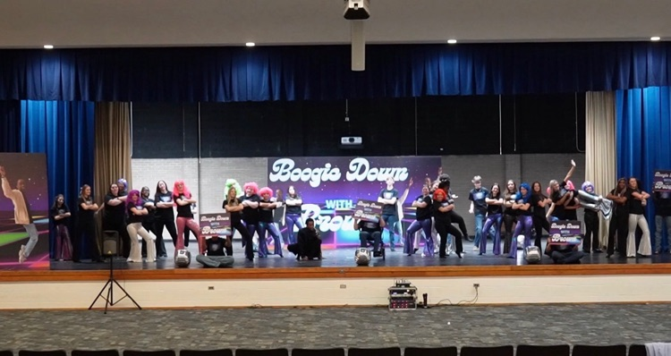 students on stage. There is a backdrop in the back that reads boogie down with Brown, students are in jeans and black pants with black T-shirt and some have colorful wigs on.