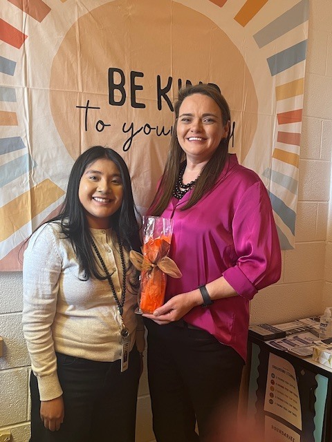 Two women stand smiling in front of a decorative wall display that reads “Be Kind to Yourself.” One woman is presenting the other with a wrapped gift, recognizing her during Social Worker Appreciation Week at Elizabethtown Middle School.