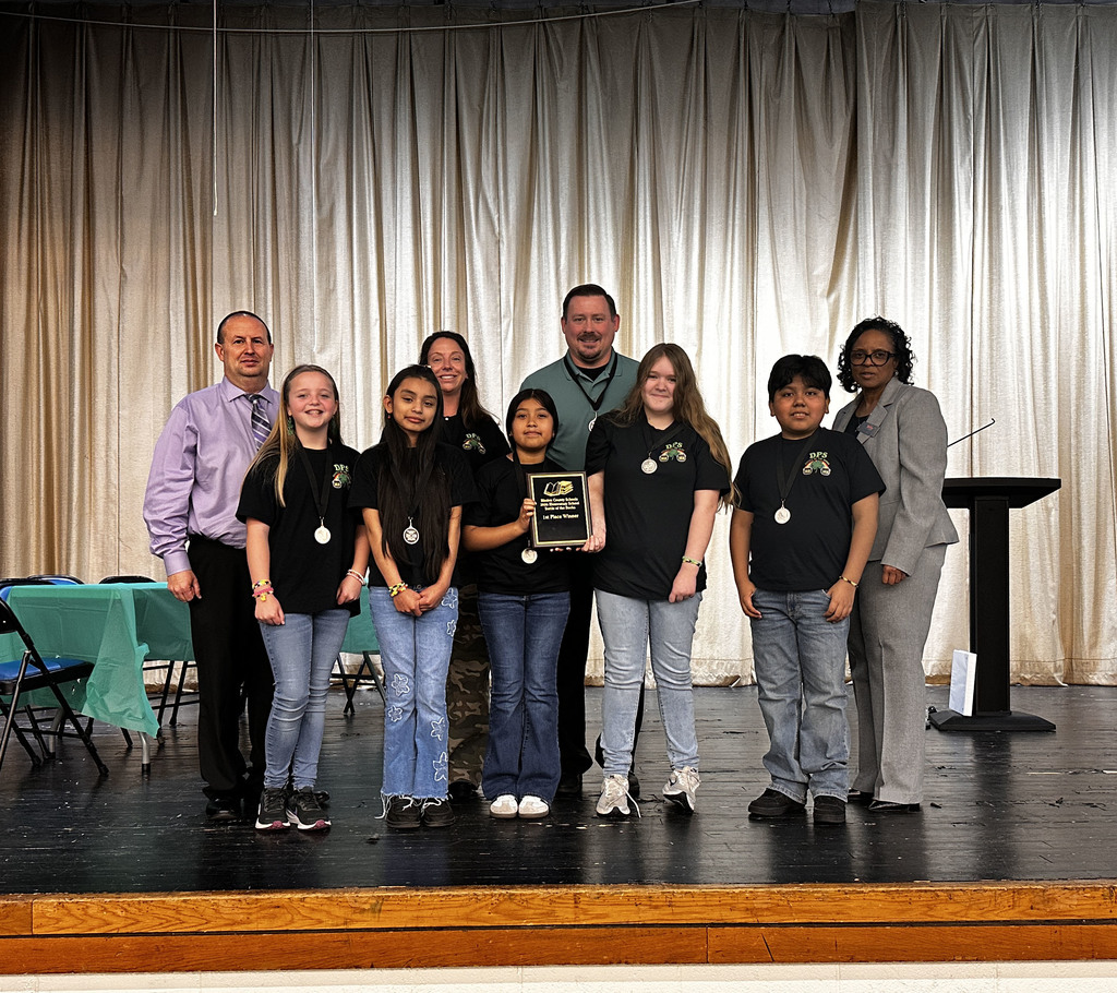 A group of elementary students wearing matching black team shirts and medals stand on a stage with several adults behind them. One student holds a plaque recognizing their team’s first-place finish in the Battle of the Books competition.