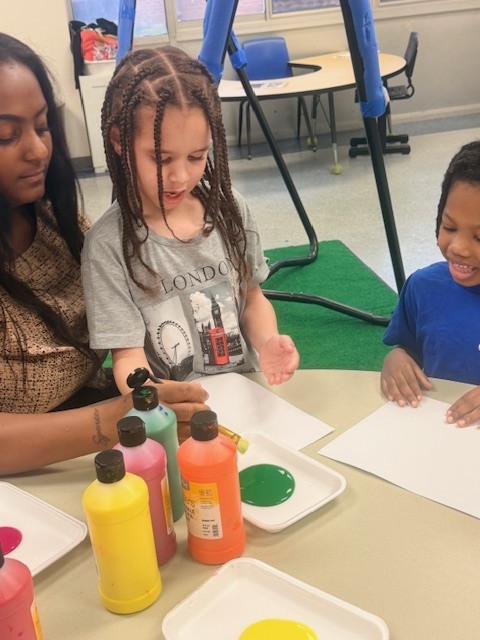 A young student smiles while painting with a teacher assisting, surrounded by bottles and trays of colorful paint during a classroom art project.