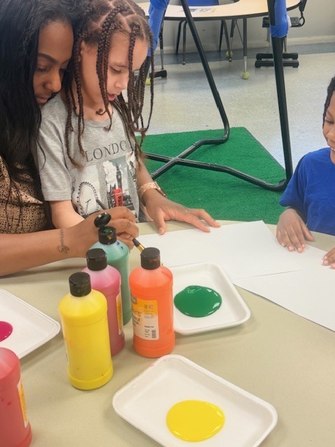 A student dips a paintbrush into bright paint while sitting beside a teacher and another student at a table with trays of yellow, green, and orange paint.