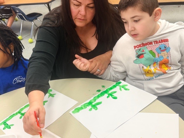 A teacher helps a student paint green stems on paper while another student watches during a classroom art activity.