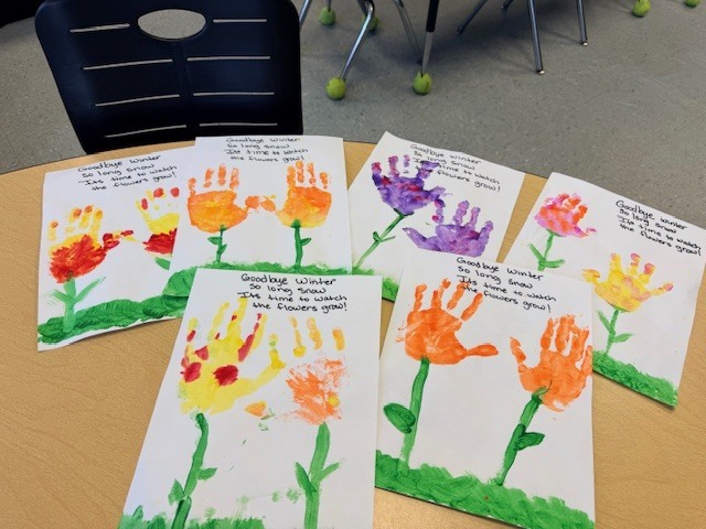 Several completed student paintings showing colorful handprint flowers with green stems and leaves, representing human-made flowers used to say goodbye to winter.