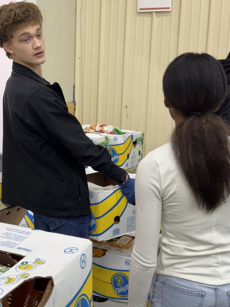 A young man in a black jacket looks toward a person with dark hair in a ponytail at a community food bank event