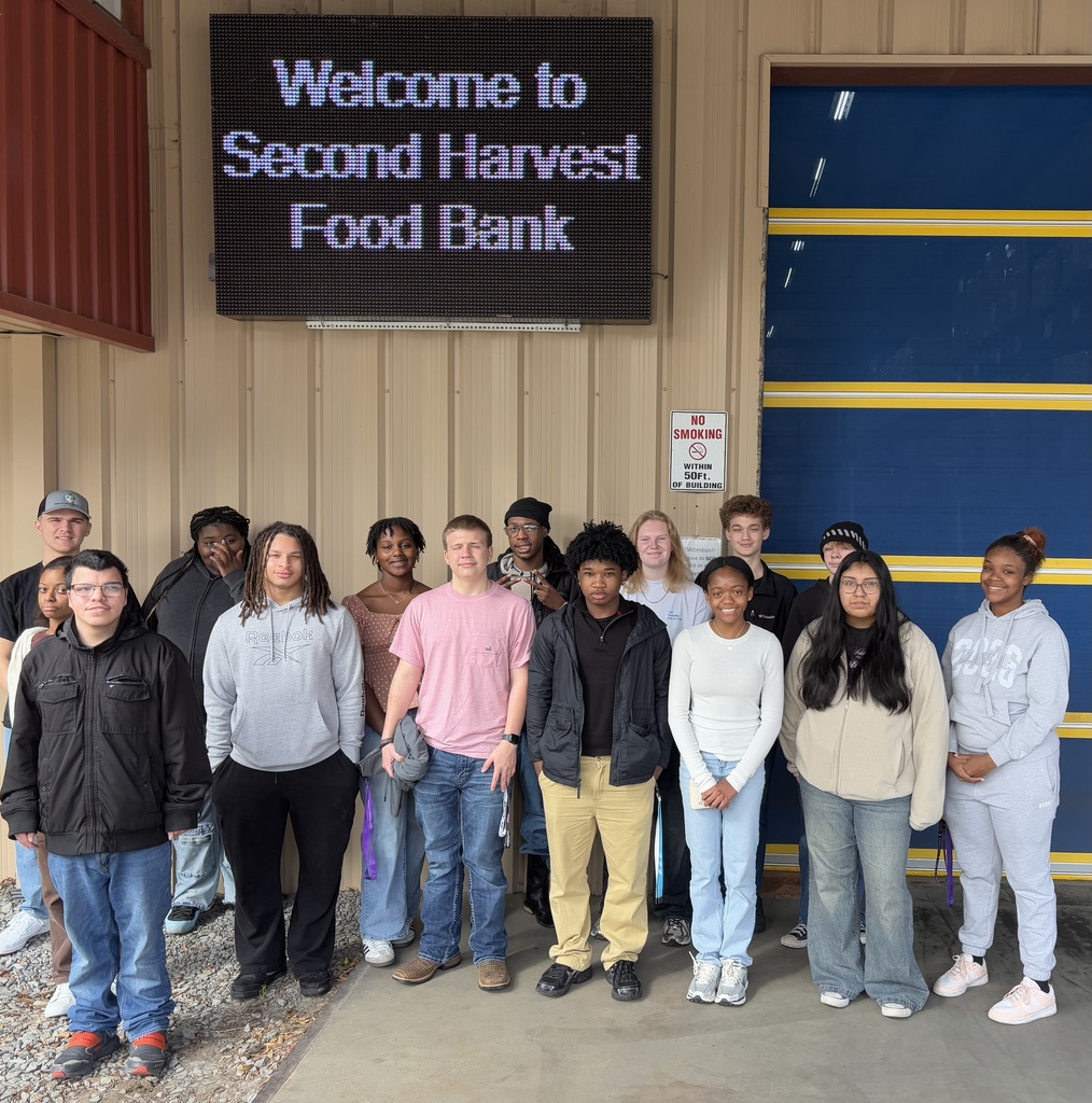A group of students stands in front of a tan building under a sign that reads "Welcome to Second Harvest Food Bank."