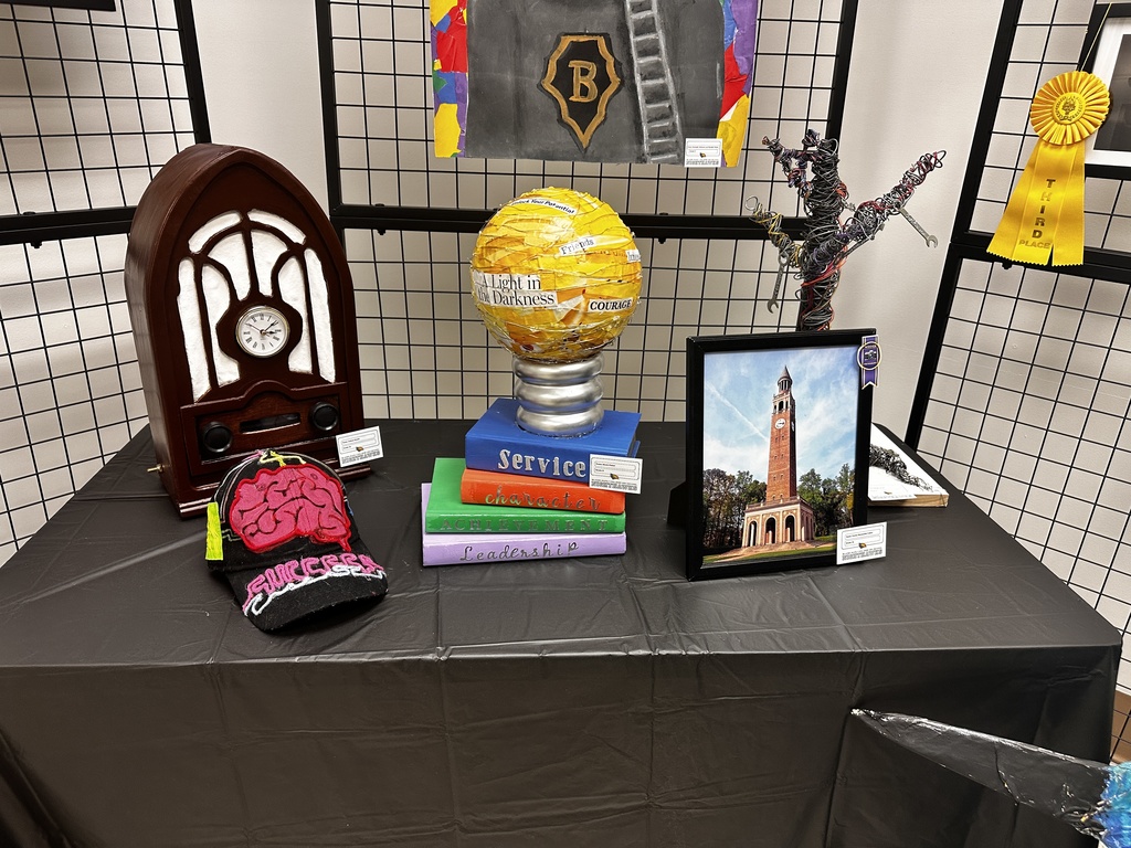Table displaying student sculptures including a vintage radio model, a decorated graduation cap with a brain design, a globe sculpture labeled with words like service and leadership, a framed photograph of a clock tower, and a wire sculpture figure.
