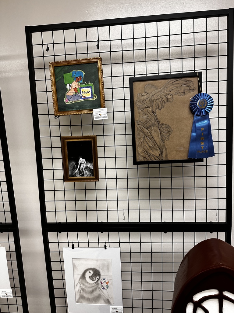 Student artwork display featuring a first-place ribbon beside a large relief drawing of a winged figure, along with a framed painting of a girl reading, a black-and-white photograph, and a pencil drawing of a penguin.