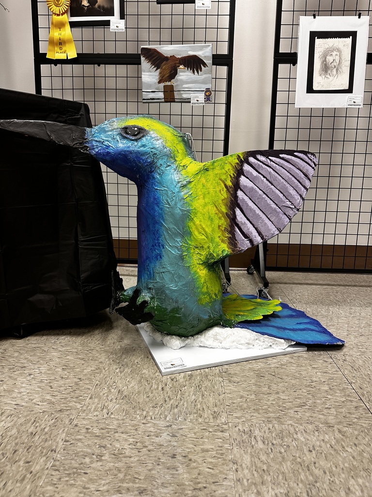 Large colorful bird sculpture with blue, green, and yellow painted body and wings, displayed on a base in front of a student art exhibition panel.
