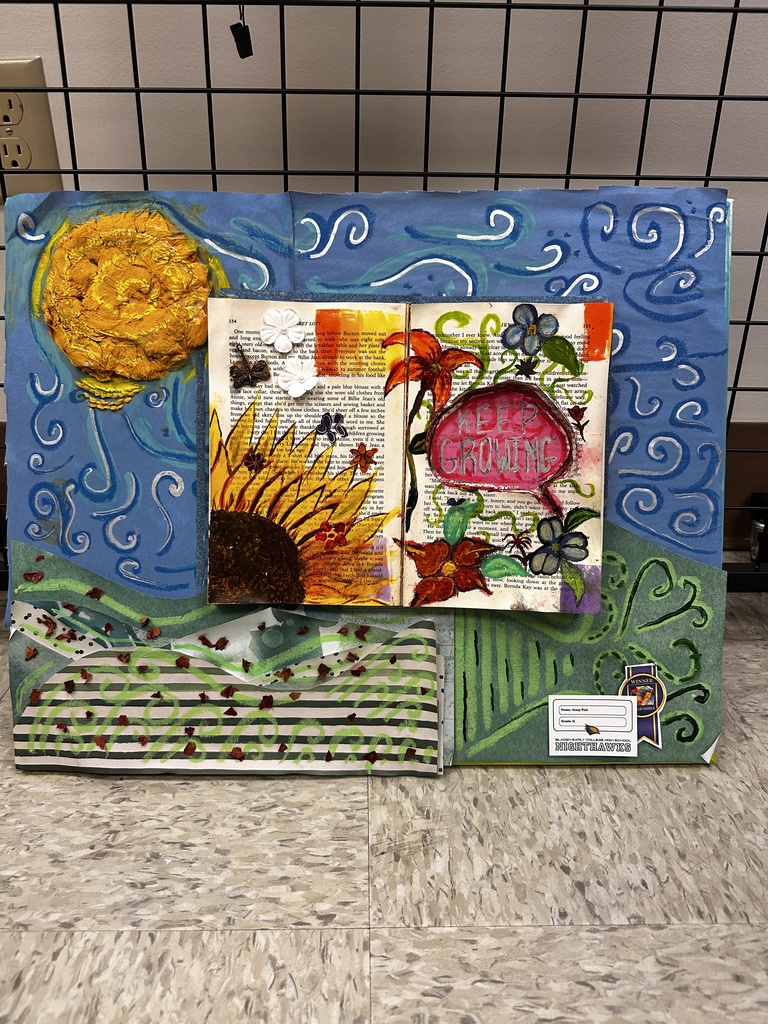 Large mixed-media artwork displayed on the floor featuring an open altered book surrounded by painted waves, flowers, and a textured yellow sun with the words “Keep Growing” on the page.