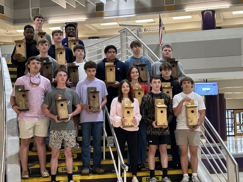 A group of approximately fifteen high school students standing on a staircase indoors, many of them proudly holding handmade wooden birdhouses. A large American flag is visible in the background.