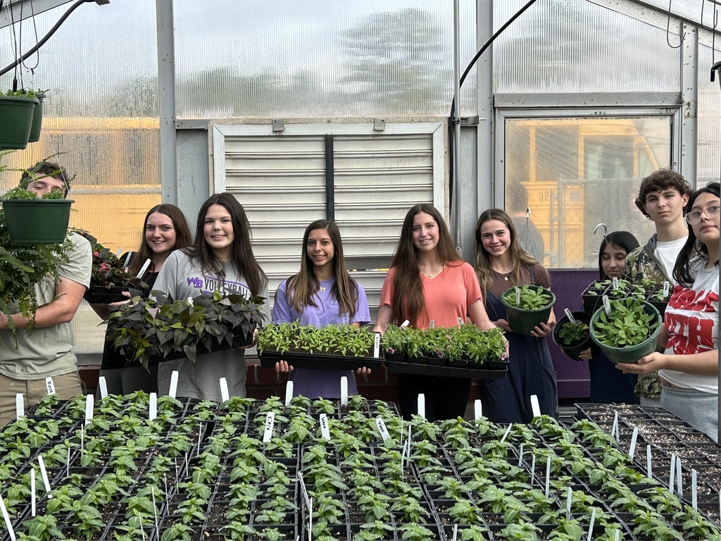 Eight smiling West Bladen High School horticulture students stand together in a greenhouse, proudly holding various potted plants and trays of green seedlings. The students are positioned behind several rows of organized plant starts, with the greenhouse glass structure visible in the background