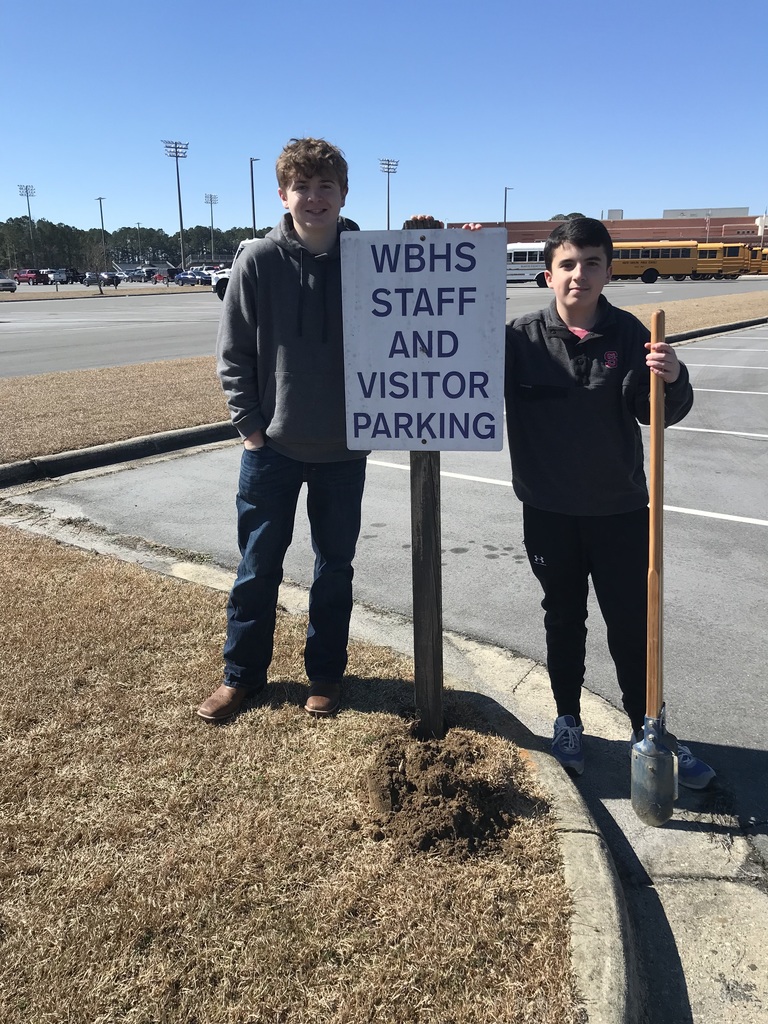 Two people standing on a sidewalk next to a patch of grass with a shovel and a hole being dug in the dirt.