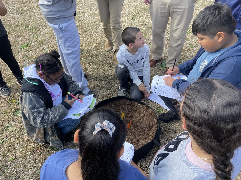 Students sit and kneel in a circle around a large container of soil outdoors while writing observations on worksheets and examining the soil together.