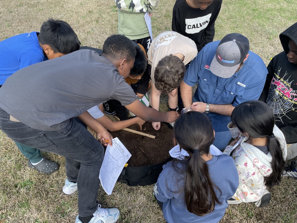A group of students gather closely around a soil container as an instructor helps them measure and examine the soil while students record notes on worksheets.