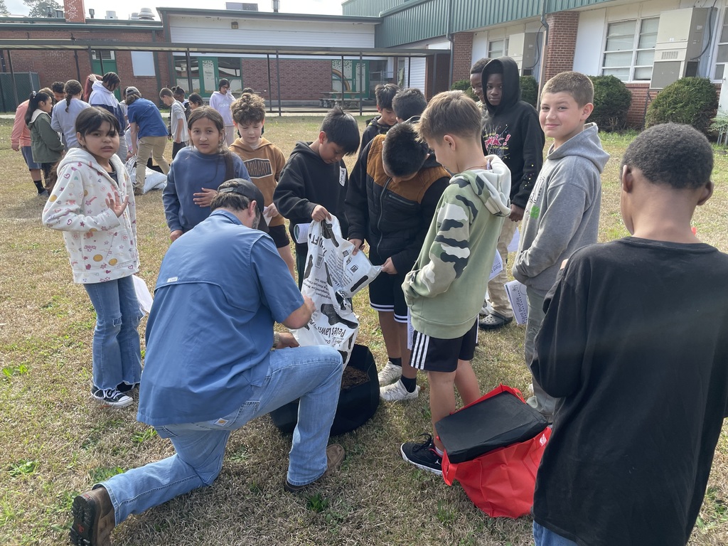 Students stand around an instructor who kneels beside a soil container and pours soil from a bag while explaining the activity.