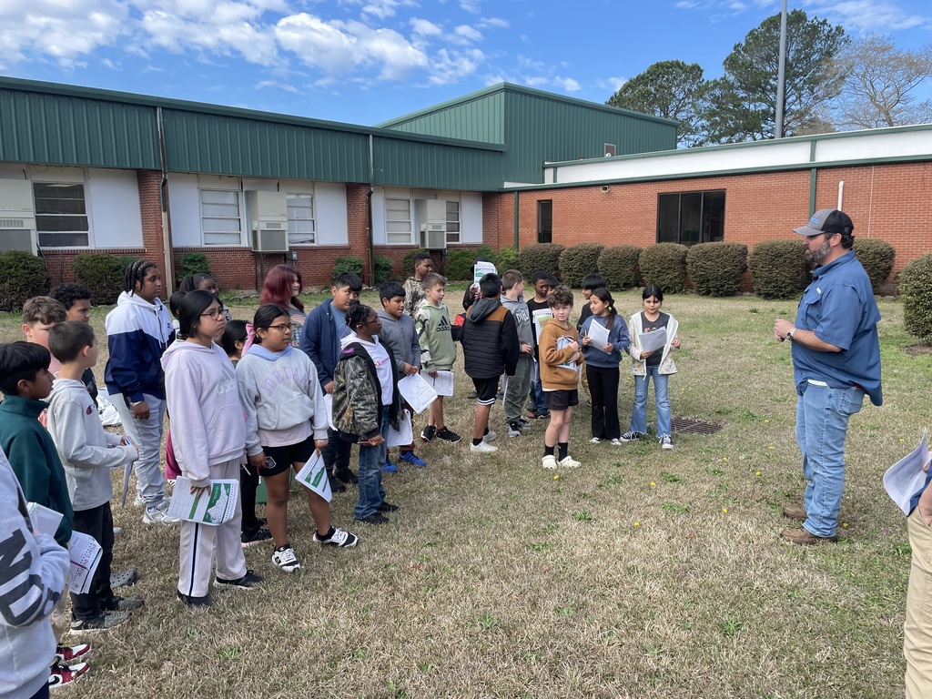 A class of students stands outside on the school lawn listening to an instructor give directions for an outdoor learning activity.