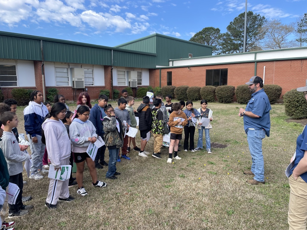 Students gather outside near their school building while listening to an instructor explain the next steps of the outdoor science lesson.