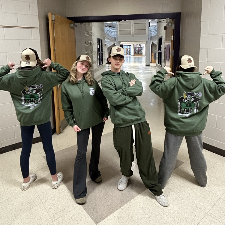 Four students in a school hallway wear matching green hoodies and tan caps. Two face forward posing, while two face away showing the hoodie logo.