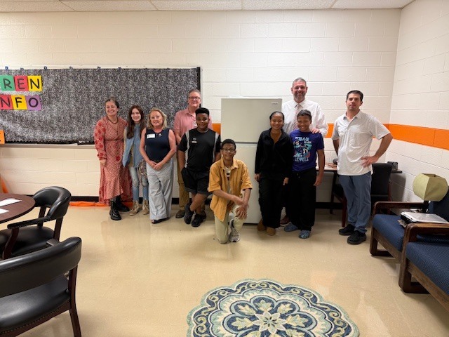 group of Elizabethtown Middle School students and adults stand together in the school’s Family Resource Center next to a newly donated refrigerator. The group poses and smiles for the photo, celebrating the Elizabethtown Rotary Club’s donation that will help expand the school’s food pantry to better serve students and families.