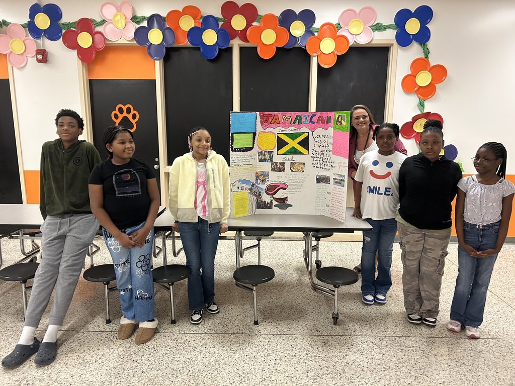 Six students and a teacher stand beside a colorful Jamaica culture display board highlighting Jamaican food, geography, and traditions during the school’s Multicultural Celebration.
