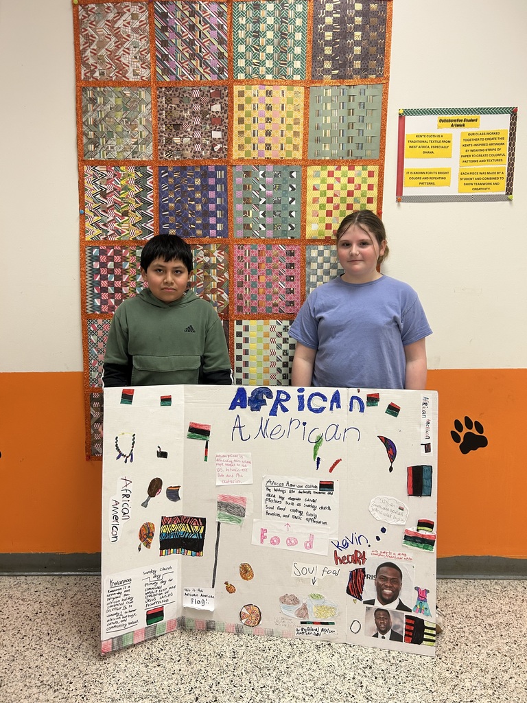 Two students stand behind a large African American culture project board, featuring information about traditions, soul food, cultural symbols, and notable figures