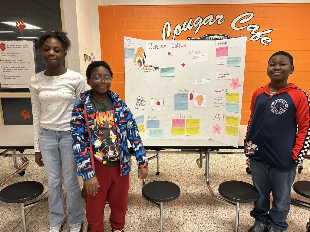 Three Elizabethtown Middle School students stand beside a tri-fold presentation board about Japanese culture, featuring information about food, lifestyle, language, traditions, hygiene, music, and art, displayed in the Cougar Café.