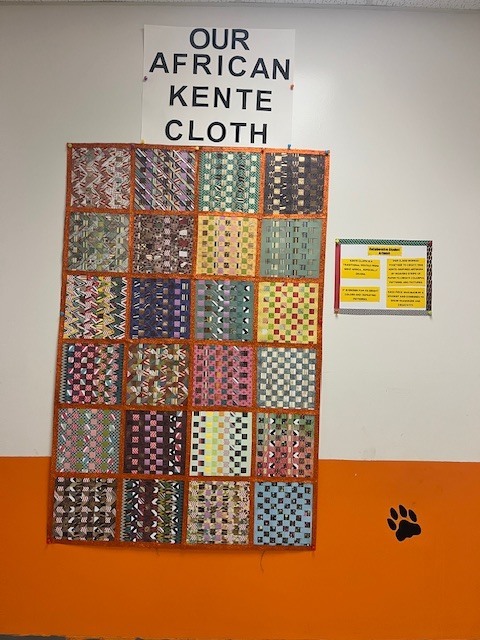 A large display titled “Our African Kente Cloth” hangs on the wall at Elizabethtown Middle School. The colorful piece is made up of many small woven squares arranged in rows, representing strips of Kente cloth created by students and staff. Each square features different patterns and colors, showing the collaborative work of Mrs. Edwards’ class during a multicultural learning project. A small informational poster explaining Kente cloth traditions is displayed beside the artwork.