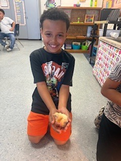 A student kneels on the classroom floor and proudly holds a baby chick while smiling at the camera.