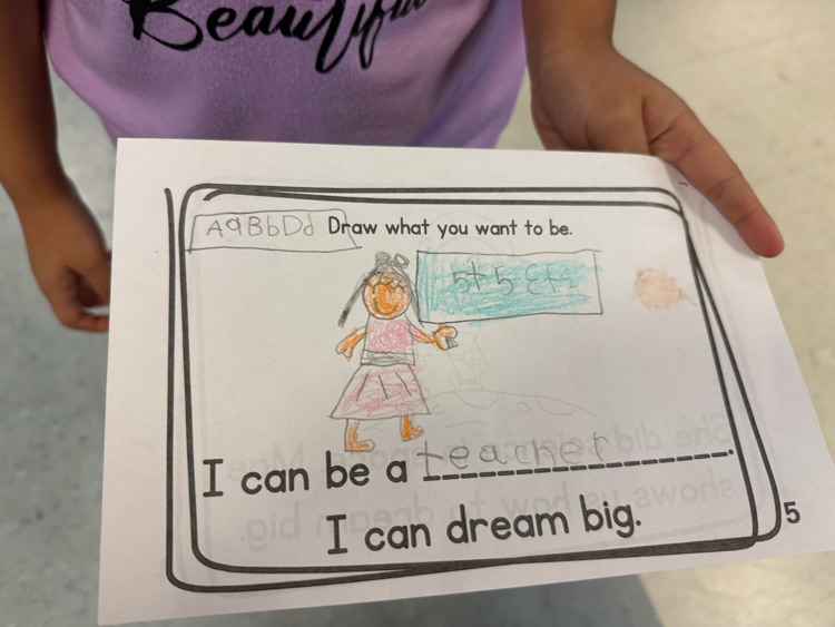 Student holding a worksheet drawing of a teacher standing beside a board that says “ABC.”