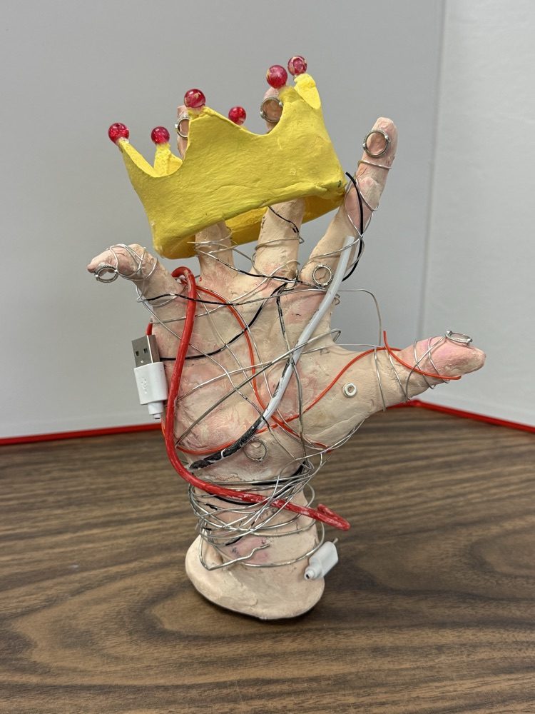Mixed-media sculpture of a hand wrapped in wires and cords with a yellow crown above it, displayed on a table