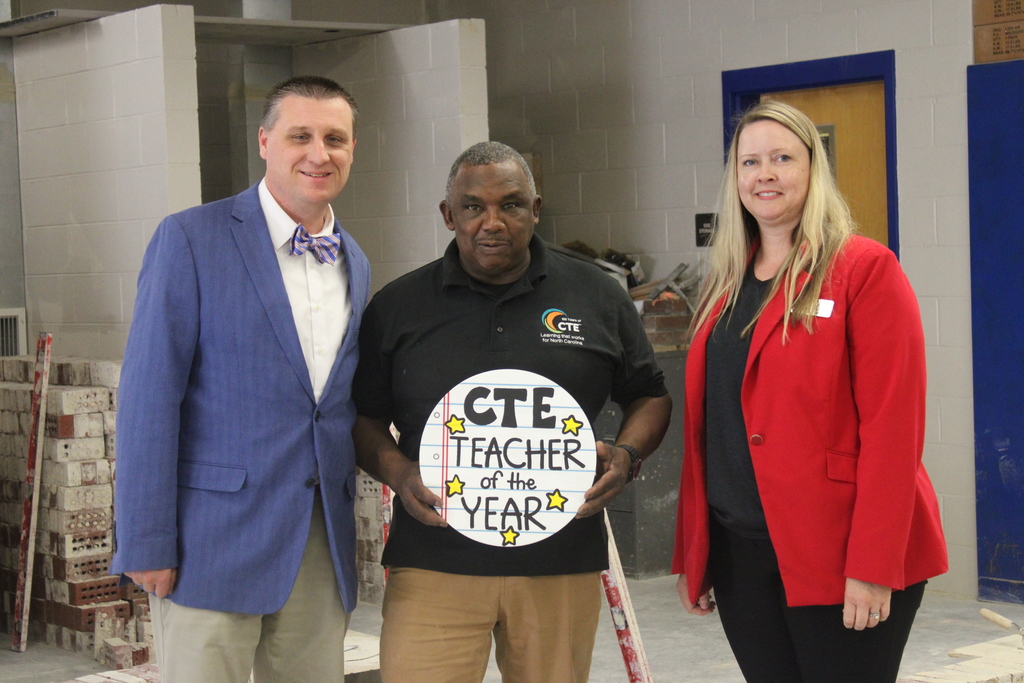Three adults stand together inside a construction training classroom. The person in the center holds a round sign that reads “CTE Teacher of the Year.” Stacks of bricks, masonry materials, and a ladder are visible behind them, suggesting a hands-on building or masonry lab environment. The two people on either side are dressed in professional attire, and all three are facing the camera and smiling.