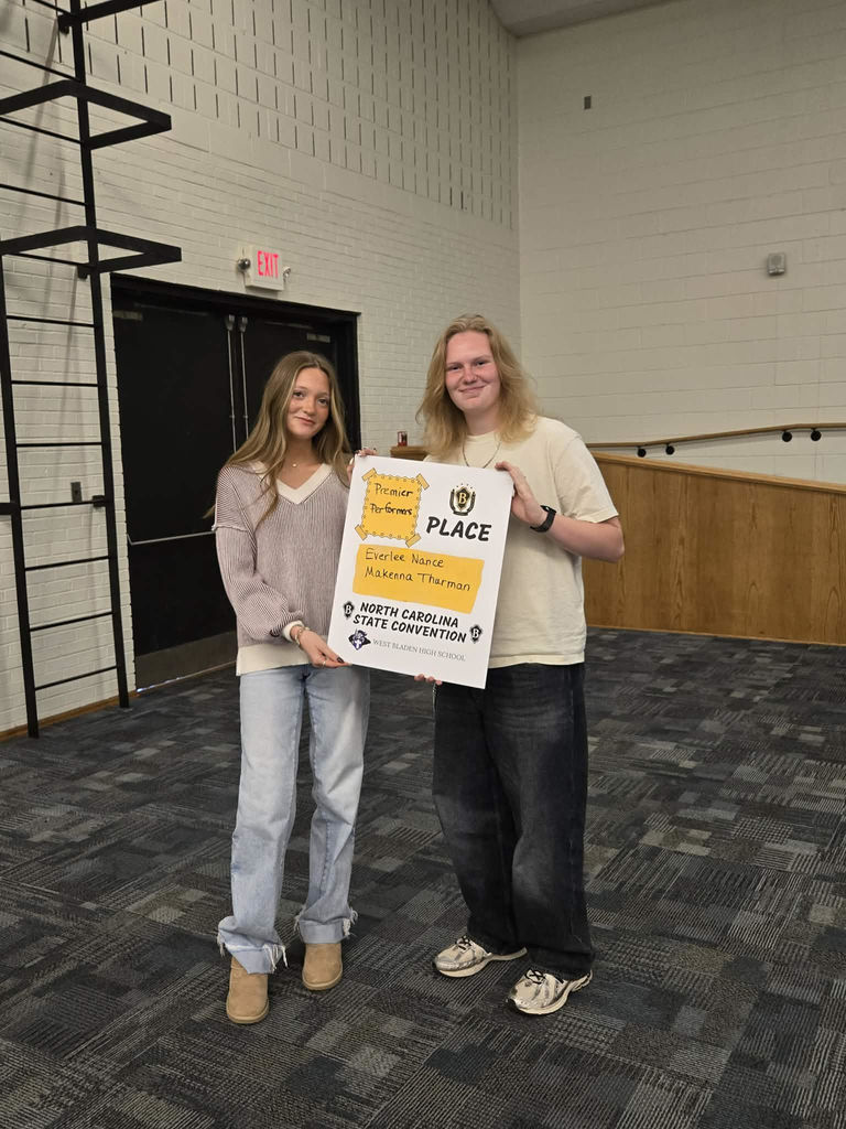 Two smiling West Bladen High School students hold a "Premier Performers" award certificate while standing inside a school building
