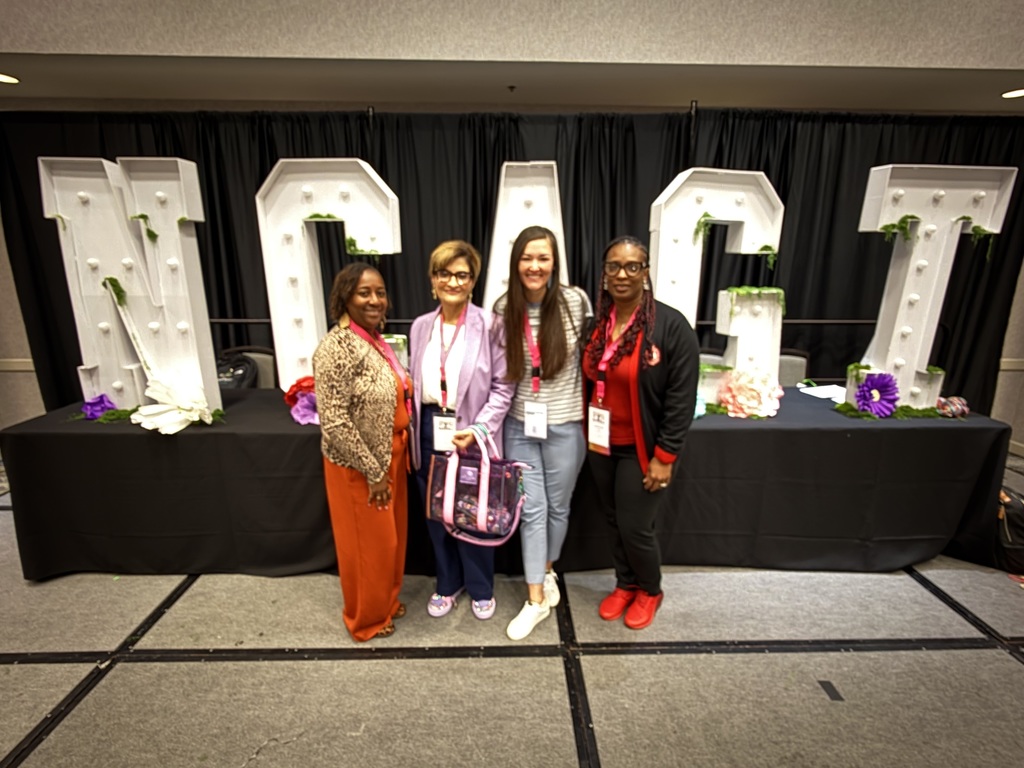Four educators pose in front of large illuminated decorative letters on a stage at a conference venue, wearing name badges and smiling during a professional learning event.