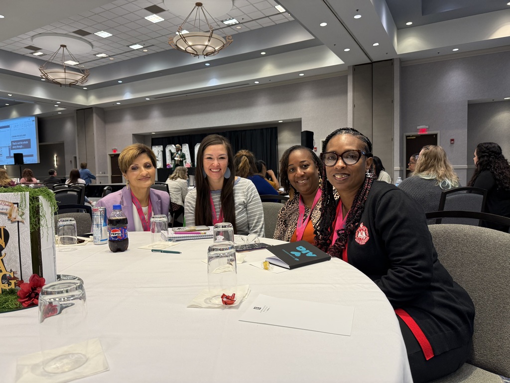 Four educators sit together at a round conference table inside a large ballroom during a professional development event, smiling toward the camera with notebooks, water glasses, and conference materials on the table.