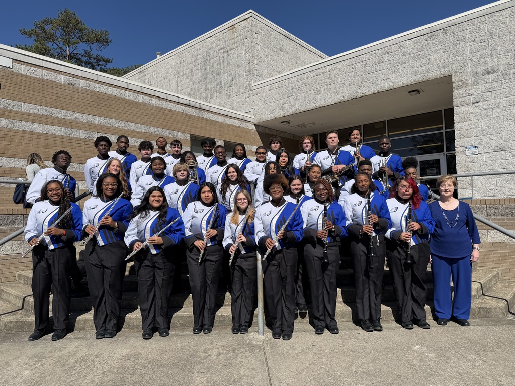 Large group of student band members in uniform pose on steps outside school holding instruments with staff nearby.